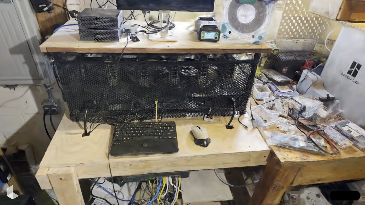 Gaussian splat capture of the GPU rig, an open-frame GPU box behind black mesh on a plywood bench in the basement, keyboard and mouse in front, cabling everywhere
