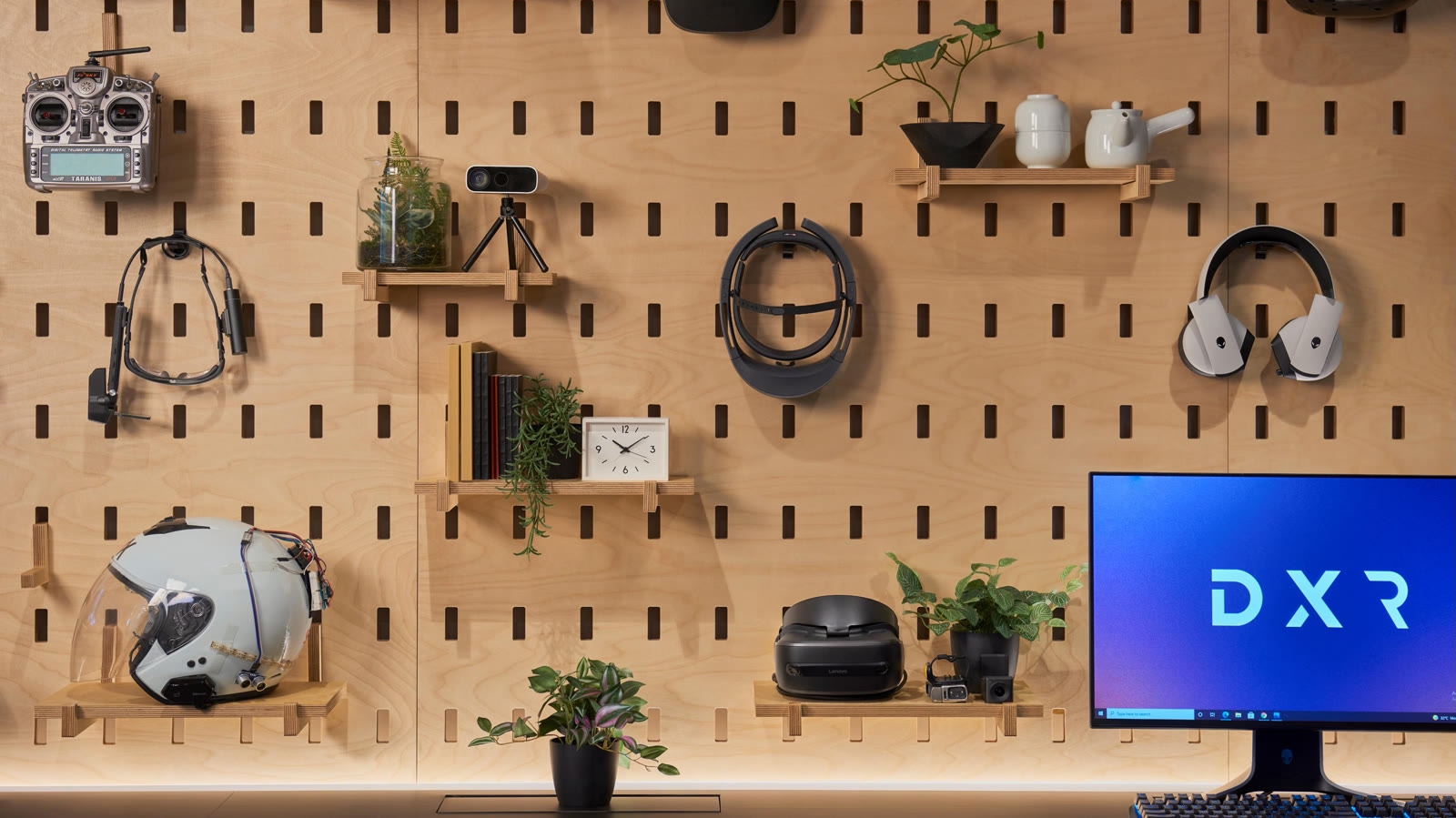 The hardware wall at Dell XR Studios, a birch pegboard hung with VR headsets, a custom AR helmet, an RC transmitter and a monitor showing the DXR logo.